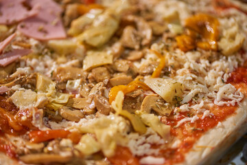 A close-up of a homemade rectangular pizza topped with ham, artichokes, cheese, and red peppers, ready for the oven.