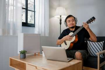 Asian man with sitting on a sofa in his home's living room, picking up a guitar.	