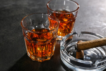 Glasses of cold rum and ash tray with cigar on black grunge background, closeup