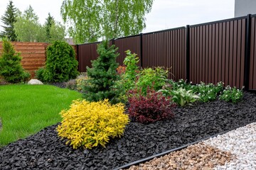 Lush garden bed with vibrant plants and black mulch against a modern brown fence creating a serene outdoor scene showcasing landscape design and horticultural beauty