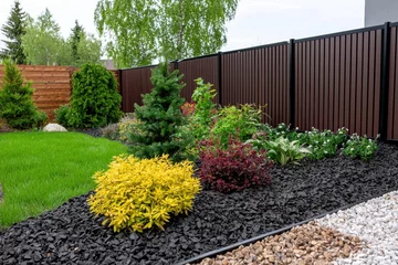 Gordijnen Lush garden bed with vibrant plants and black mulch against a modern brown fence creating a serene outdoor scene showcasing landscape design and horticultural beauty © Faizal