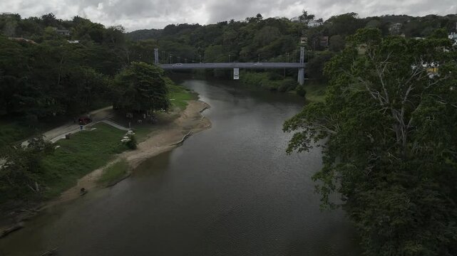 Drone lowers over Macal RIver on cloudy afternoon with metal suspension bridge crossing the river in San Ignacio, Belize