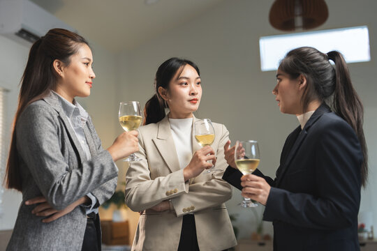 Three professional women converse while holding glasses of white wine. They appear to be networking or discussing business in a relaxed, social setting. The atmosphere is friendly and engaging