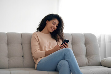 Obraz premium Happy young Hispanic woman uses smartphone at home. Smiling female scrolls social media shops online. Relaxed on comfy couch. Casual and modern wear. 3d illustration