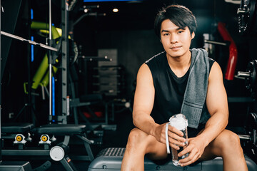 Portrait of Asian young man trained body in black sportswear sitting for rest holding water or protine in fitness gym, Weight training, healthy lifestyle concept