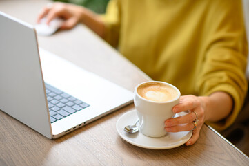 A woman works on her laptop in a café, sipping coffee. The cozy atmosphere and soft lighting create a productive yet relaxed environment for her to focus and enjoy her beverage.