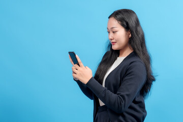 Naklejka premium A focused woman wearing a dark sweater stands and interacts with a smartphone against a bright blue studio backdrop.