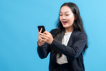 Happy businesswoman holding smartphone, looking at screen with excited expression, standing against blue background. Concept of technology, communication, success, and online news.