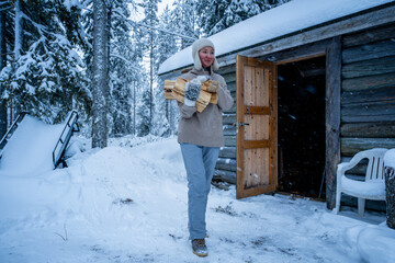 Woman in warm clothes carrying firewood outside a rustic log cabin