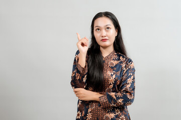 A young woman in stylish Indonesian batik attire, pointing upwards with a thoughtful expression,...