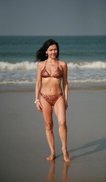 Pretty woman 51 years old in a fashionable swimsuit with sequins stands on the beach against the backdrop of ocean waves Goa India