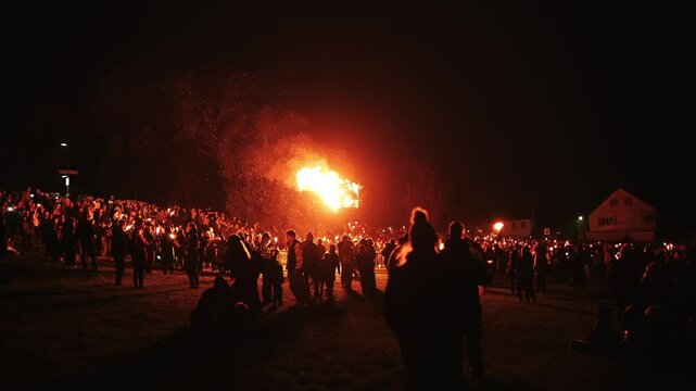 A large crowd of people are gathered around a fire, with some holding lit candles. The atmosphere is lively and festive, with people enjoying the warmth and light of the fire