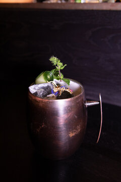 A stylish cocktail served in a rustic metal mug, garnished with cucumber, fresh herbs, ice, and edible flowers on a dark surface.