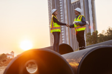 Two engineers shake hands on large water pipes at a construction site, celebrating the completion...