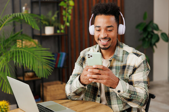 Young man of African American ethnicity wears casual clothes listen to music in headphones use mobile cell phone sitting at table in coffee shop cafe relax rest in restaurant during free time indoors.
