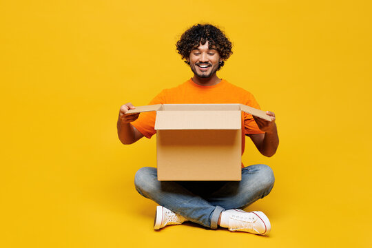 Full body smiling cheerful fun happy young Indian man he wear orange t-shirt casual clothes sit open unpack blank cardboard box isolated on plain yellow background studio portrait. Lifestyle concept.