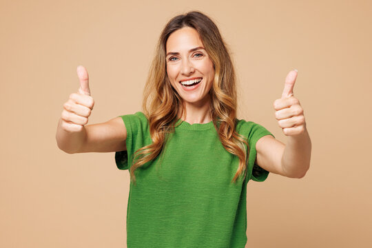Young smiling happy cheerful fun cool woman she wearing green t-shirt casual clothes showing thumb up like gesture isolated on plain pastel light beige background studio portrait. Lifestyle concept.