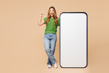 Full body young woman wear green t-shirt casual clothes big huge blank screen mobile cell phone smartphone with workspace area point index finger up isolated on plain pastel light beige background.