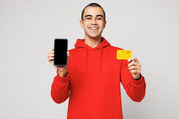 Young middle eastern man wear red hoody casual clothes use blank screen mobile cell phone hold credit bank card shopping online order delivery booking tour isolated on plain solid white background.