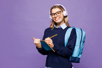 Young girl student wear blue casual clothes glasses headphones backpack bag taking notes write down...
