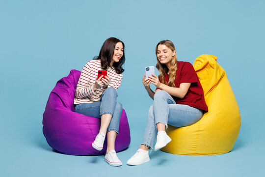 Full body young couple two friends women wearing red casual clothes together sit in bag chair hold in hand use show mobile cell phone isolated on pastel plain light blue background. Lifestyle concept. - Powered by Adobe