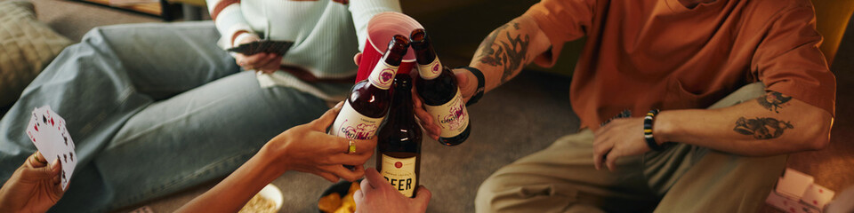 Friends sitting together, playing cards and raising beer bottles for toast during leisure activity....