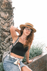 A young woman in a stylish hat stands near tall palm trees on a sunny day. A young tourist walking on the streets of Europe.