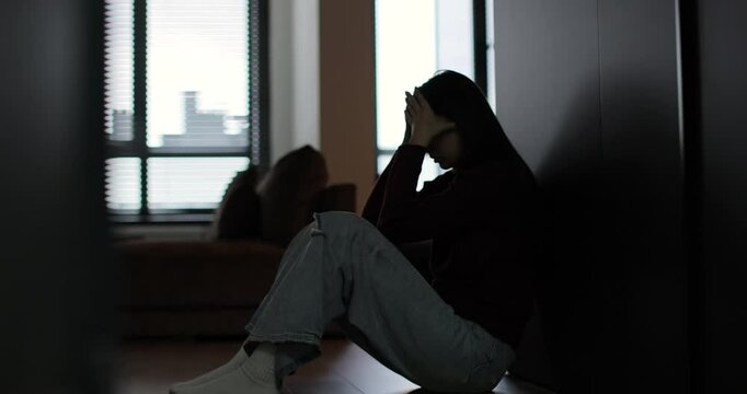 Silhouette of a sad woman sitting in the dark leaning against the wall in home. Domestic violence, family problems, stress, violence.