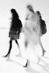 The blurred motion of people walking is shot in black and white. The composition focuses on their blurred shapes against an isolated background, emphasizing modern fashion with sharp lines and strong 
