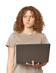 Young Caucasian redhead woman with laptop confused, feels doubtful and unsure.