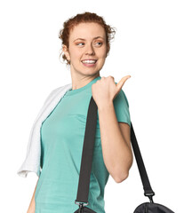 Young Caucasian redhead with gym gear points with thumb finger away, laughing and carefree.