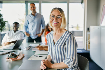 Smiling businesswoman Leading Diverse Meeting in Modern Office. She leans over the table as she looks into the camera and smiles. Her three diverse colleagues look smilingly in the same direction