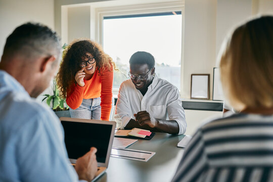 Diverse team collaborating in a modern office environment. They sit and stand around a table while talking and smiling. On the table there is a laptop, documents and notebooks