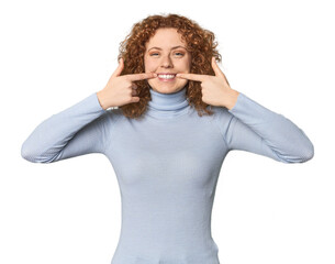 Young Caucasian redhead woman smiles, pointing fingers at mouth.