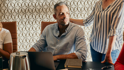 Business Meeting With Diverse Team Engaged Around Modern Office Table. The man is sitting in front of a laptop wearing a blue shirt. A female colleague, standing behind him, looking at the computer