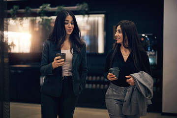 Smiling businesswomen walking with coffees through a hotel lobby. They talk and smile about a successful meeting
