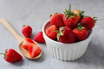 Strawberries in a white bowl with sliced stawberry in wooden spoon on grey cement background