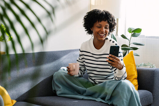 Young happy woman using mobile phone while enjoying hot tea or coffee drink relaxing on the sofa in a cozy winter day at home