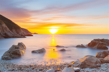 Naklejka premium A breathtaking coastal scene at sunset, with the sun setting behind jagged rocks and illuminating the vibrant ocean water with warm golden hues.