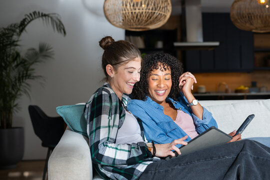 Sitting on couch, lesbian couple are smiling and looking at tablet together