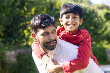 indian father giving piggyback ride to young son, both smiling outdoors