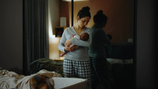 Authentic footage of a mother in pajamas at night trying to put her newborn baby to sleep in her arms. The mother is very tired and yawns and cuddles her baby in the bedroom