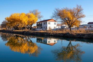 Fototapeta premium Autumn trees mirroring on river, house in background, clear sky, scenic location