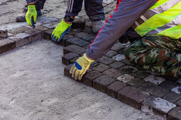 Workers laying cobblestone pavement, close-up of hands in protective gloves placing bricks.