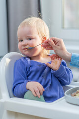 Cute baby refuses to eat when mom feeds her with a spoon in the kitchen