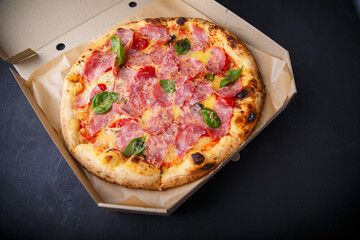 Pizza with cheese, ham, green basil leaves and tomatoes in paper box on black background. Food product advertising banner, copy space