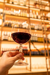 A young woman holds a fine crystal glass of wine in the foreground with a wine cellar from Tuscany on a large unfocused wood shelves. Concept of tasting, wine library, delicacy and sophistication