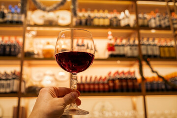 the hand of a manholds a fine crystal glass of wine in the foreground with a wine cellar from Tuscany on a large unfocused iron shelves. Concept of tasting, wine library, delicacy and sophistication