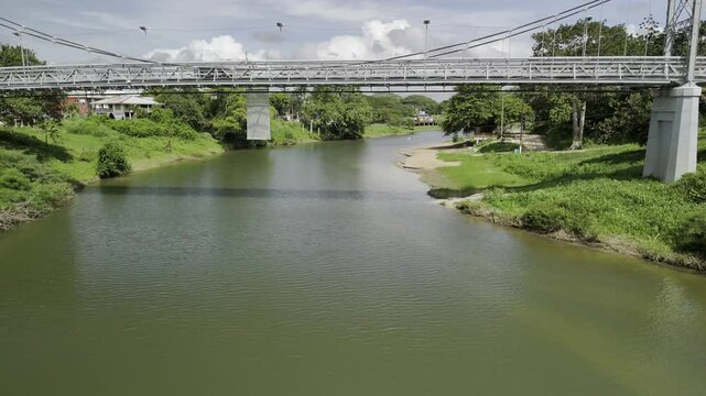 Drone facing north rises up from the Macal River to above the metal suspension Hawkesworth Bridge and overlooks San Ignacio, Belize on sunny afternoon