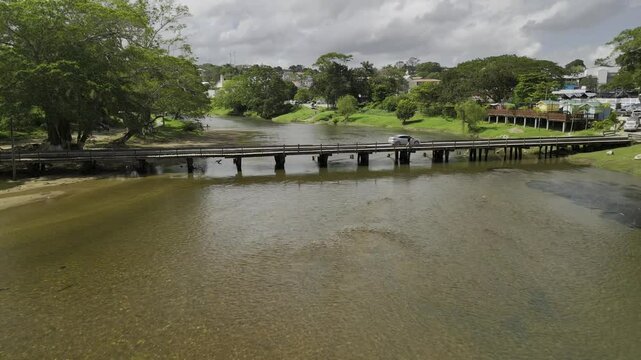 Drone flies south along the Macal River on sunny day, flying over a wooden bridge, and then under a metal suspension bridge in San Ignacio, Belize